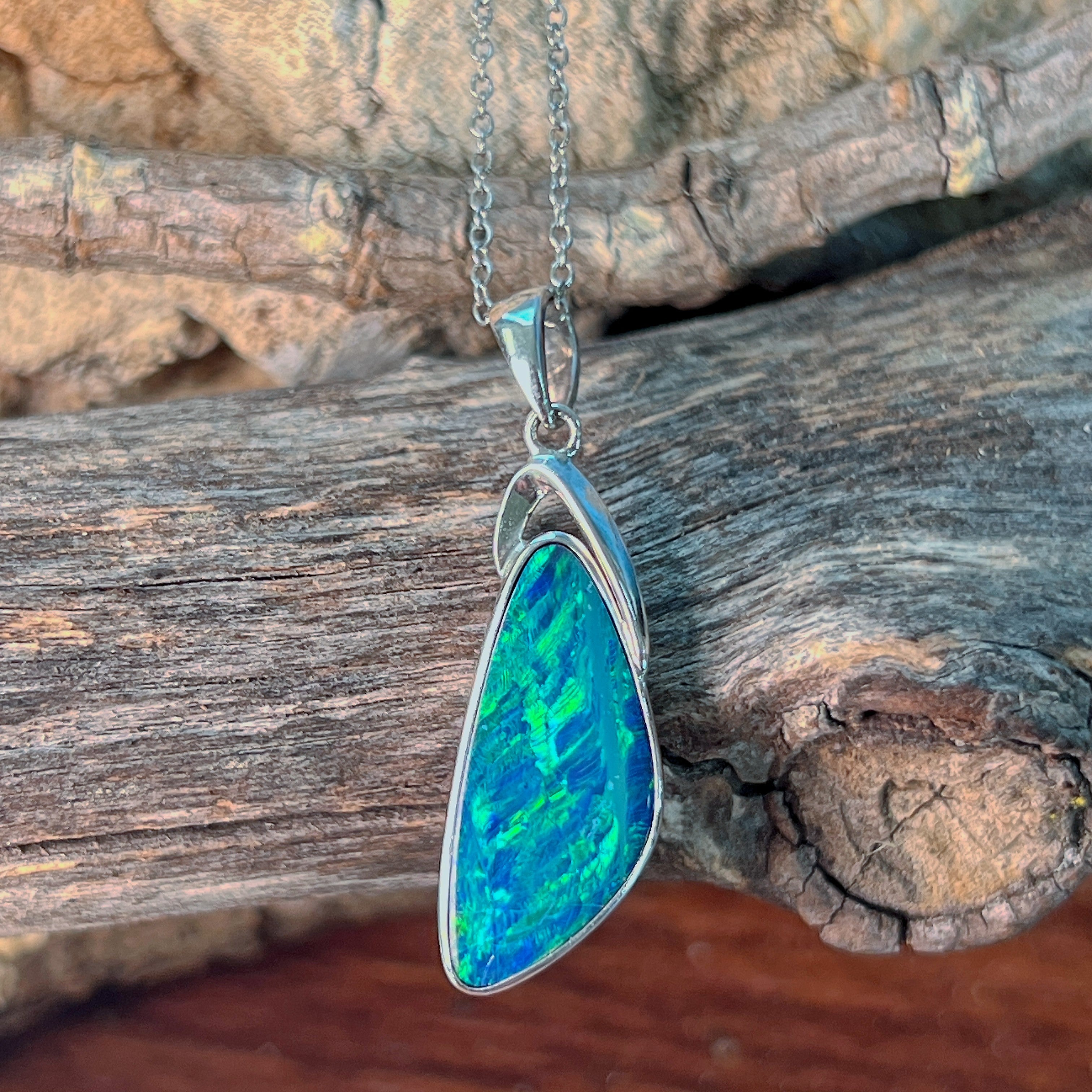Freeform triangle opal doublet pendant in silver angled against driftwood and stone