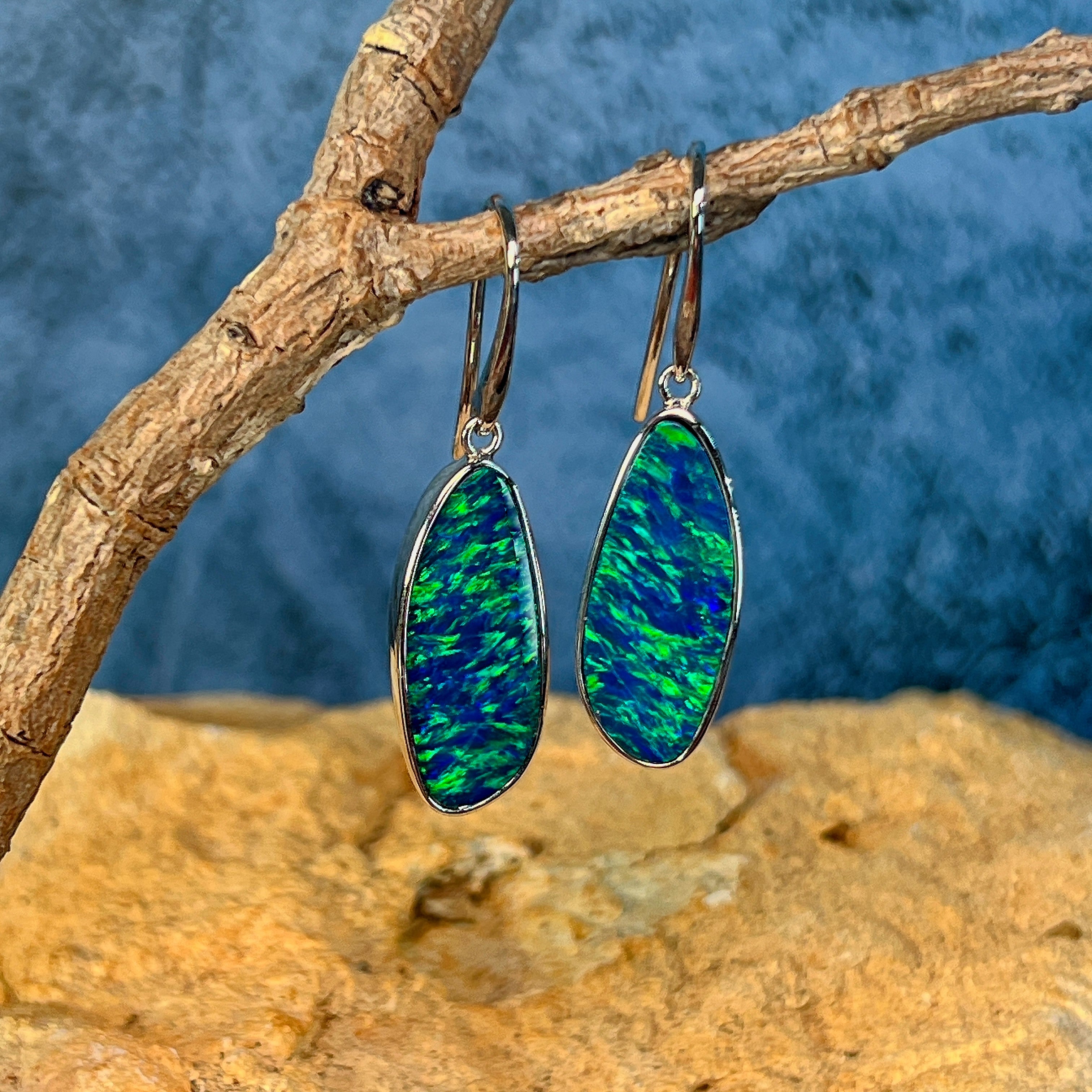 White gold opal doublet drop earrings hanging from a branch above sandstone