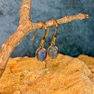 Angled view of yellow gold plated dangling earrings with 8x6mm oval opal triplets on a driftwood branch